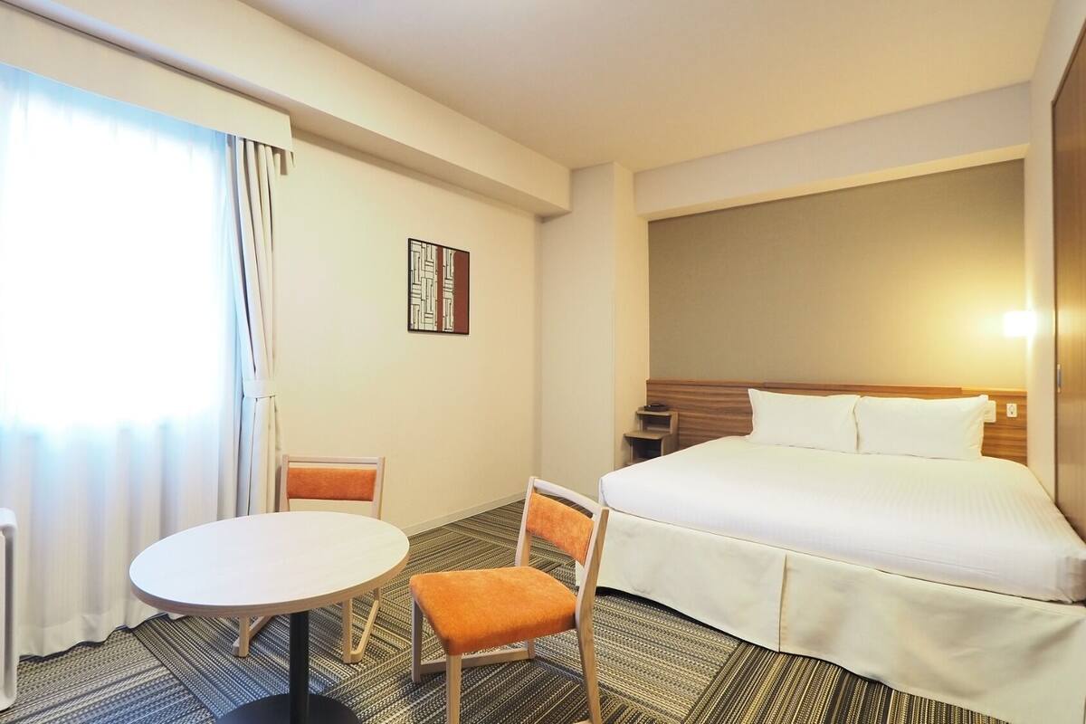 Ein Hotelzimmer mit Bett, rundem Tisch, Stuhl, Heizkörper und einem Fenster mit Vorhängen.