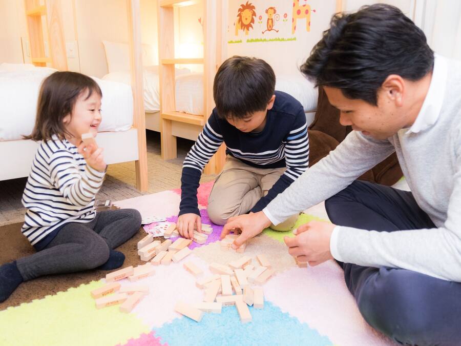 Un niño y dos adultos jugando con bloques de madera sobre una alfombra colorida en una habitación con una cama y una pared con dibujos.