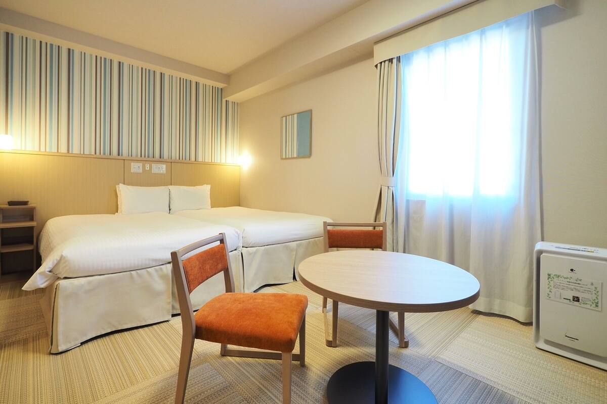 Ein Hotelzimmer mit zwei Betten, einem Sessel, einem Tisch, einer Lampe, einem Nachttisch, einem Fenster mit Vorhängen und einer Wand-Klimaanlage.