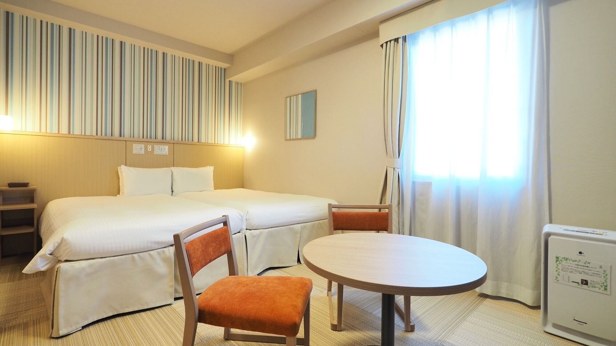 A hotel room with a bed, two chairs, a round table, a bedside table, a lamp, a mirror, and a window with curtains.