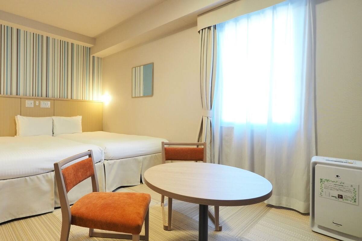 Ein Hotelzimmer mit einem Bett, zwei Stühlen, einem runden Tisch, einem Nachttisch, einer Lampe, einem Spiegel und einem Fenster mit Vorhängen.