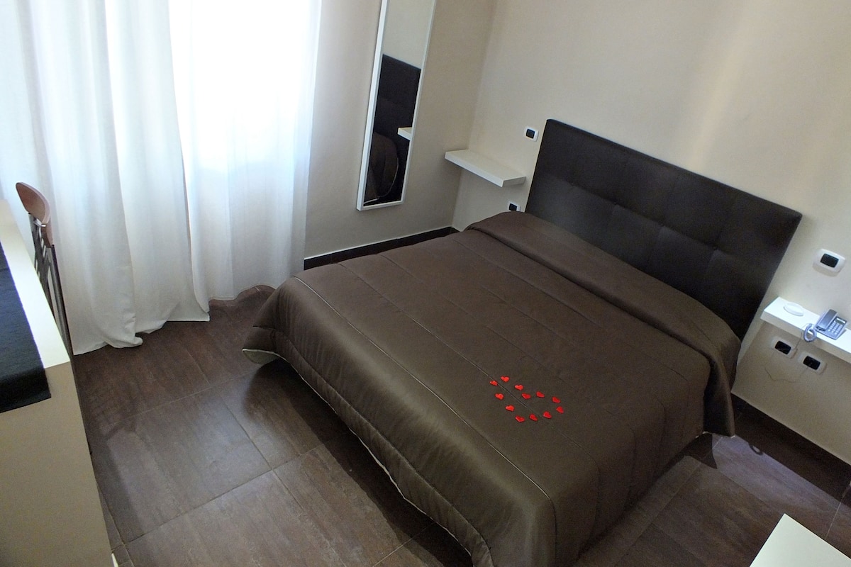 A hotel room with a bed, a bedside table, a mirror, and a window with curtains.
