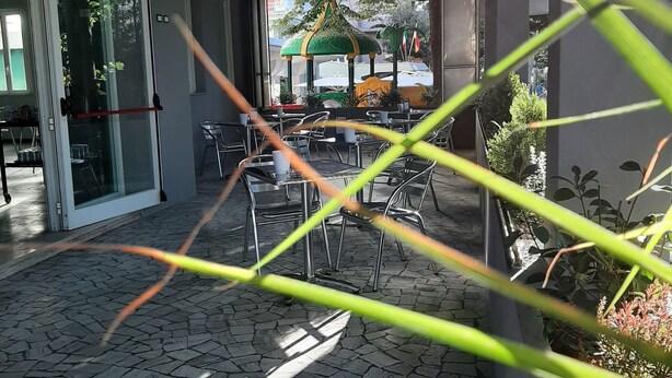 An outdoor seating area with tables and chairs, a covered section, and a view of a green-roofed structure in the background. - Hotel Alabama (Rimini)