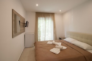 A hotel room with a bed, a TV mounted on the wall, a small bedside table, and a window with curtains. - Pension Avocado (Skiathos)