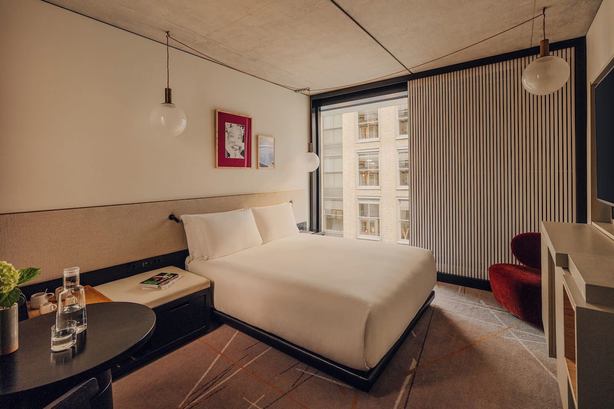 A modern hotel room with a large bed, a desk, a chair, and a view of the city through the window.
