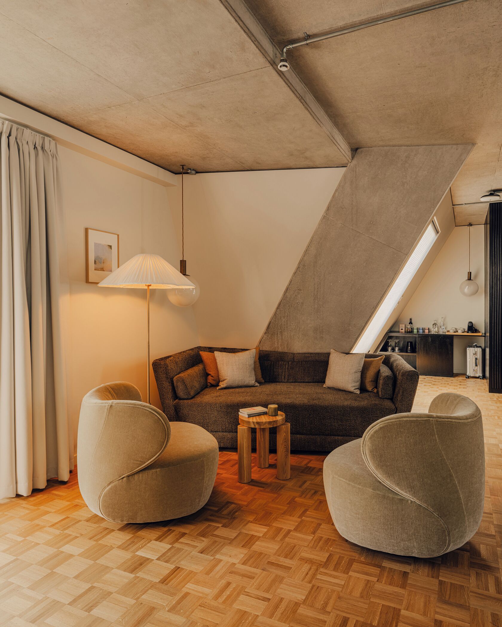 A modern loft-style living room with a herringbone-patterned wooden floor, a dark brown sectional sofa, two round armchairs, a small wooden coffee table, and a floor lamp.
