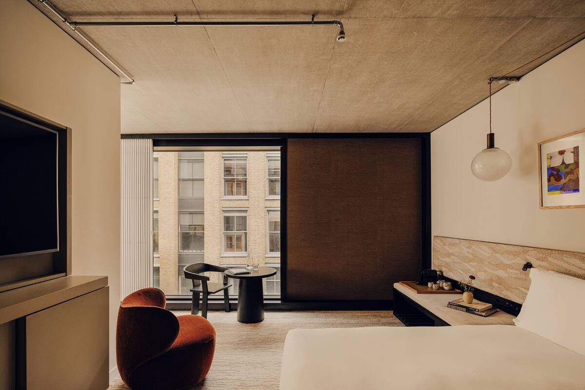 A modern hotel room with a large bed, a TV, a round table with chairs, and a view of a building through the window.