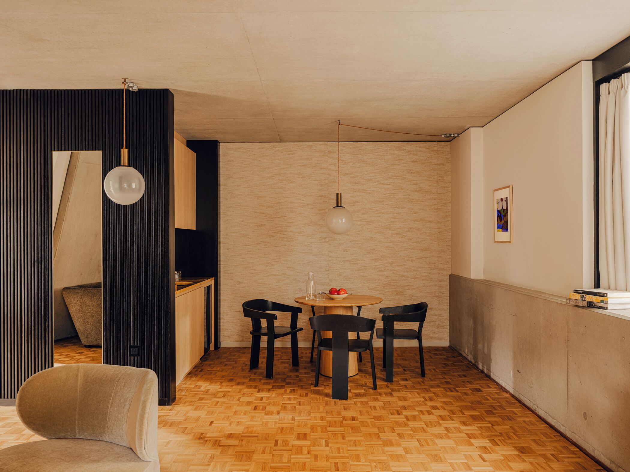 A modern living space with a wooden herringbone floor, a round wooden table with four chairs, a sofa, and a dark partition with a hanging light fixture.