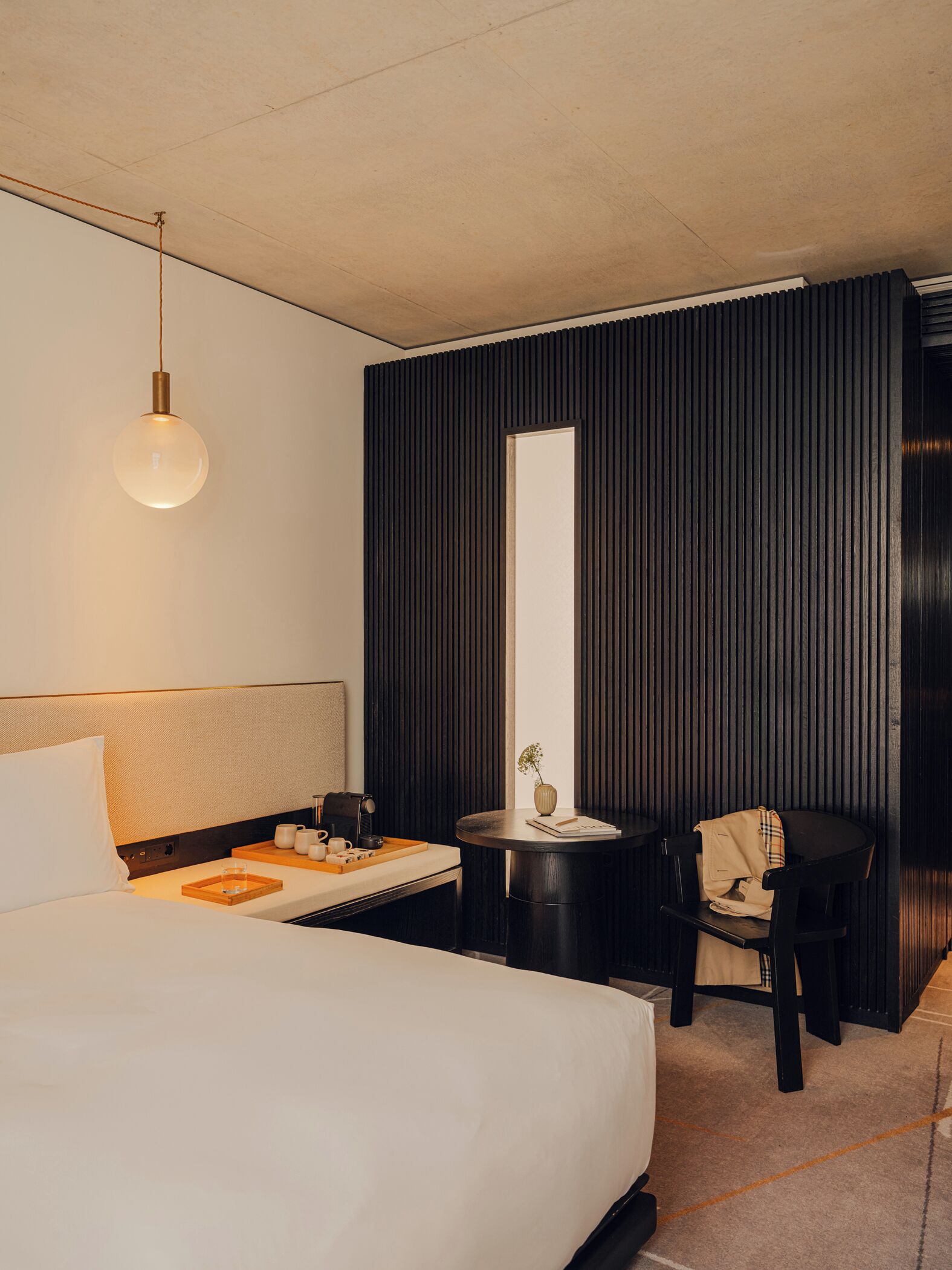 Habitación de hotel con una cama grande, una mesita de noche con lámpara, una pequeña mesa redonda con un florero y una silla.