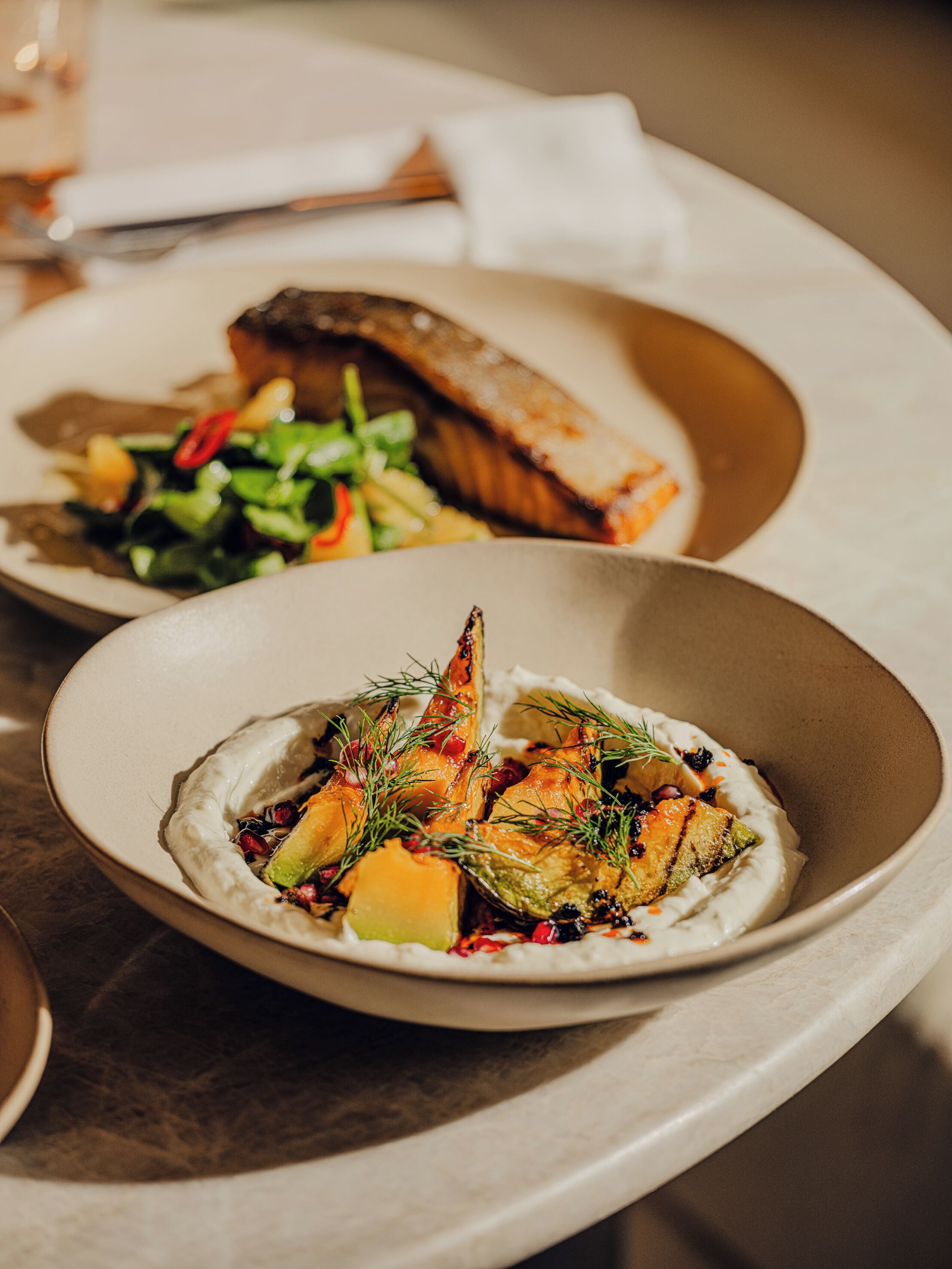A dining table with two dishes, one with a fish and vegetables and another with a creamy base and vegetables.