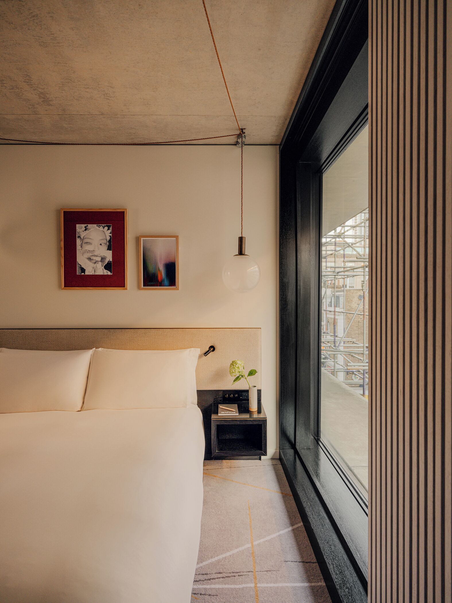 A modern hotel room with a large bed, a bedside table with a vase, and a view of the outside through a large window.