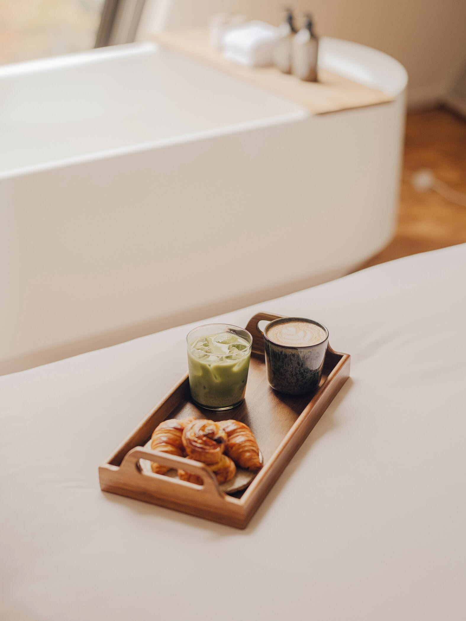 A tray with a croissant, a green beverage, and a dark coffee on a white surface near a bathtub.