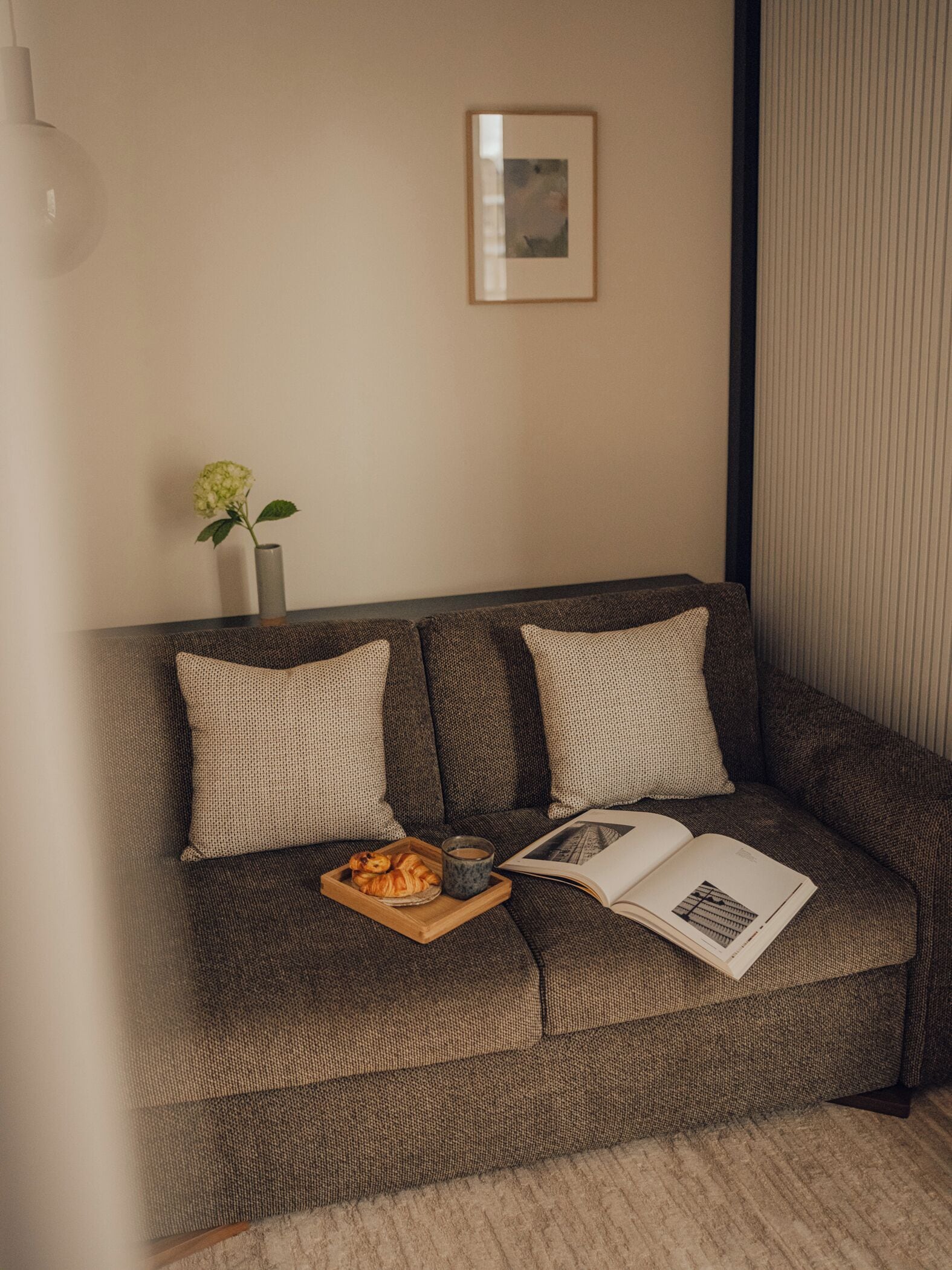 A cozy living room with a brown sofa, a small table with a book, a glass of water, and a tray of croissants.