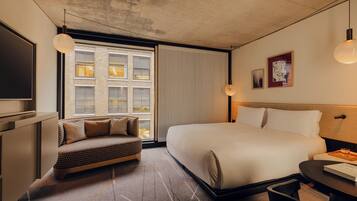 A modern hotel room with a large bed, a sofa, a TV, and a window with blinds.