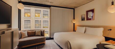 Une chambre d’hôtel moderne avec un grand lit, un canapé, une télévision et une fenêtre équipée de stores.