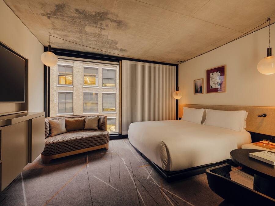 A modern hotel room with a large bed, a sofa, a TV, and a window with blinds.