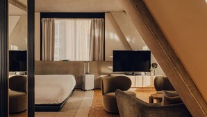 Una habitación de hotel moderna con una cama grande, un televisor de pantalla plana, una zona de estar con un sofá y sillones, y un piso de madera con patrón de espiga.