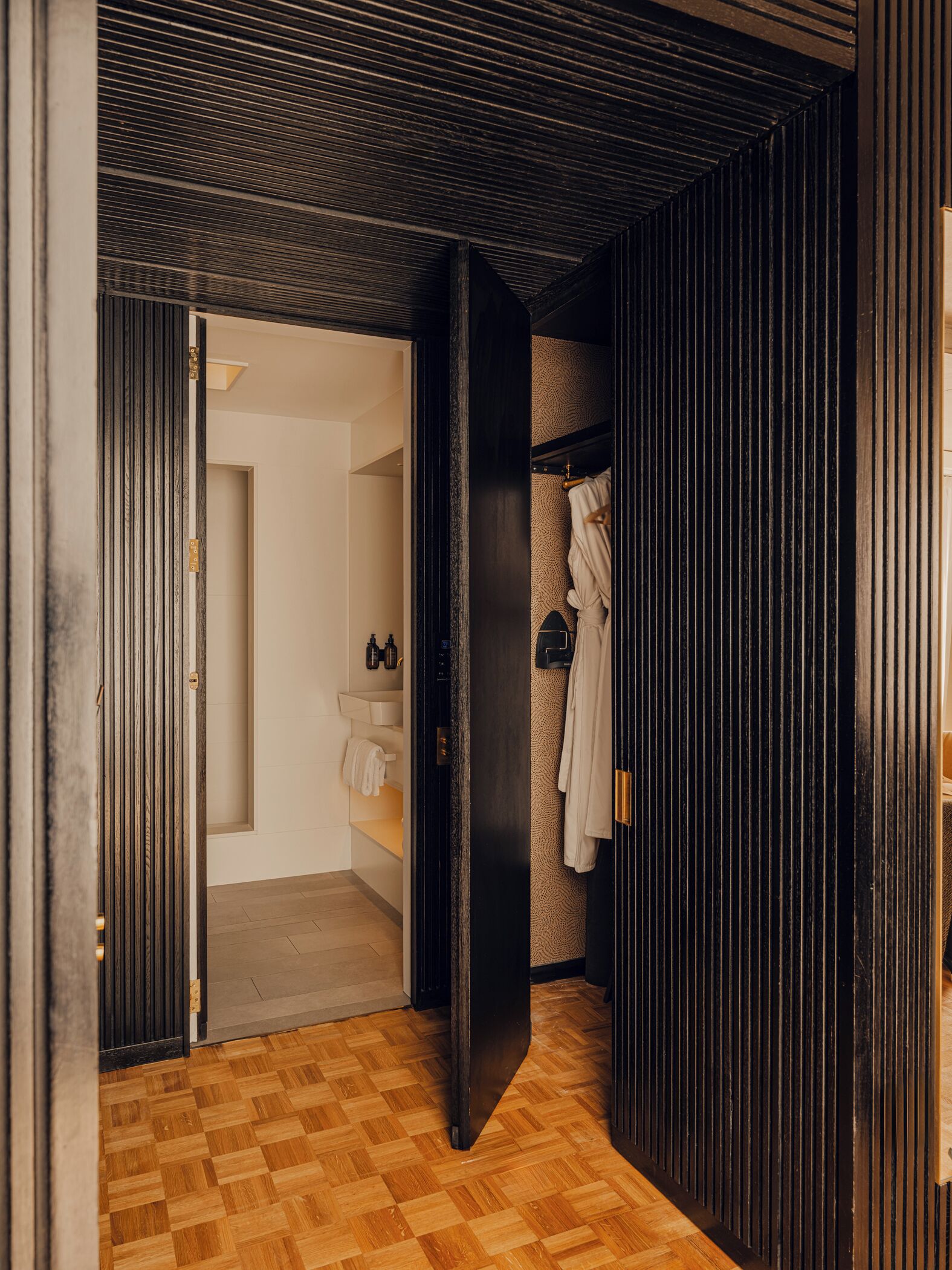 A hallway with a wooden floor leading to a bathroom with a towel rack and a sink.