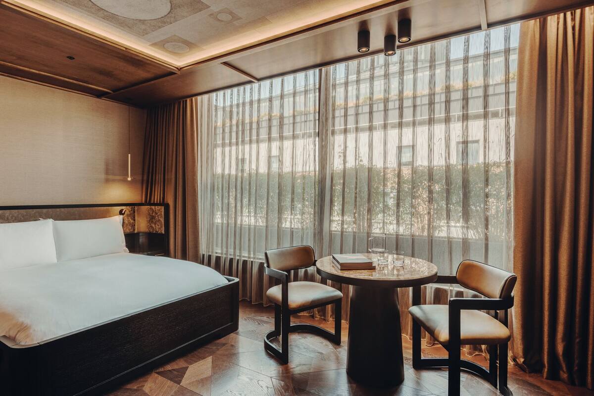 A hotel room with a large bed, two chairs, a small table, and a view through sheer curtains.