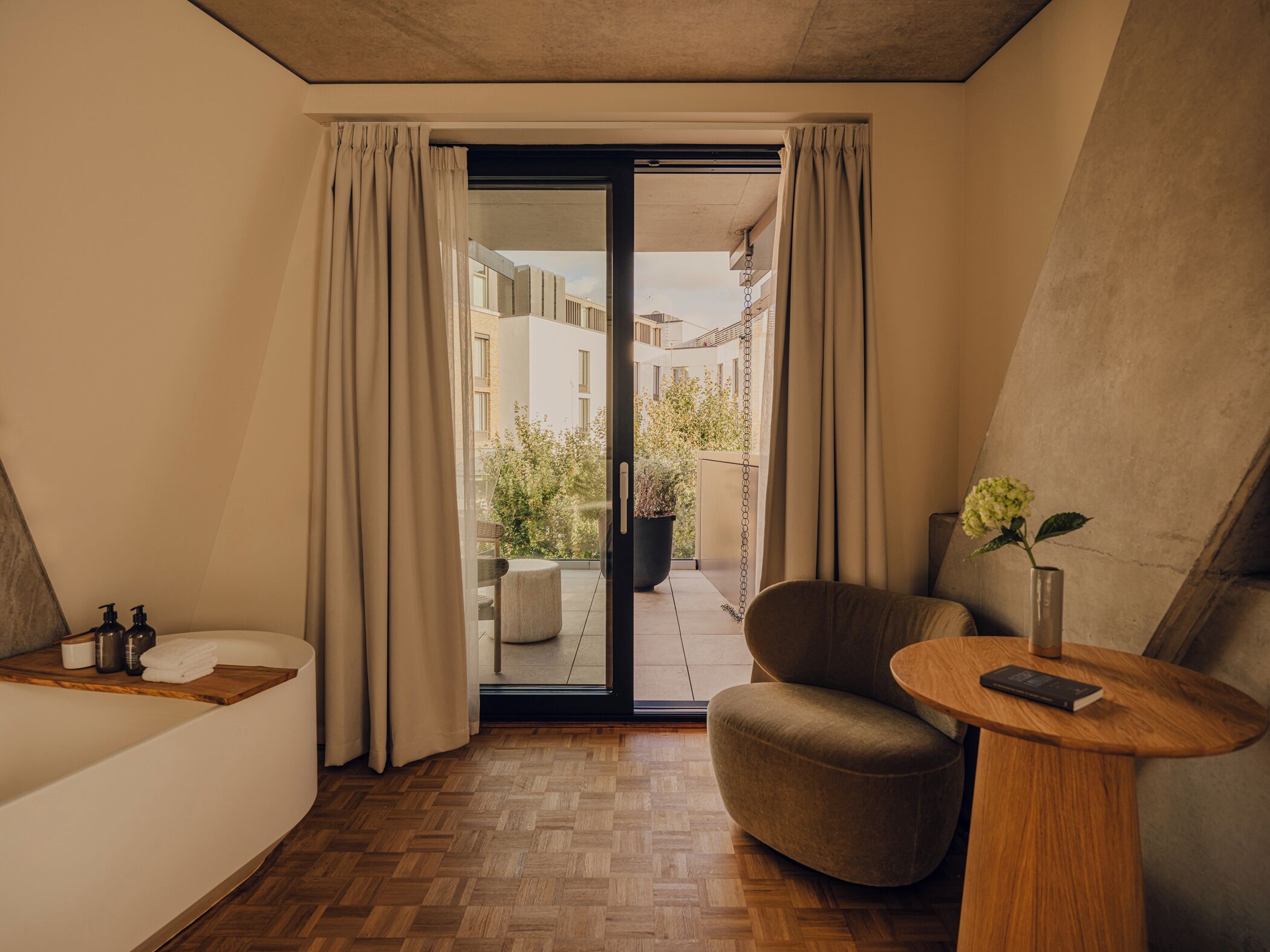 A modern hotel room with a wooden floor, a comfortable chair, a small wooden table, a potted plant, and a view of the outdoor area through a glass door.