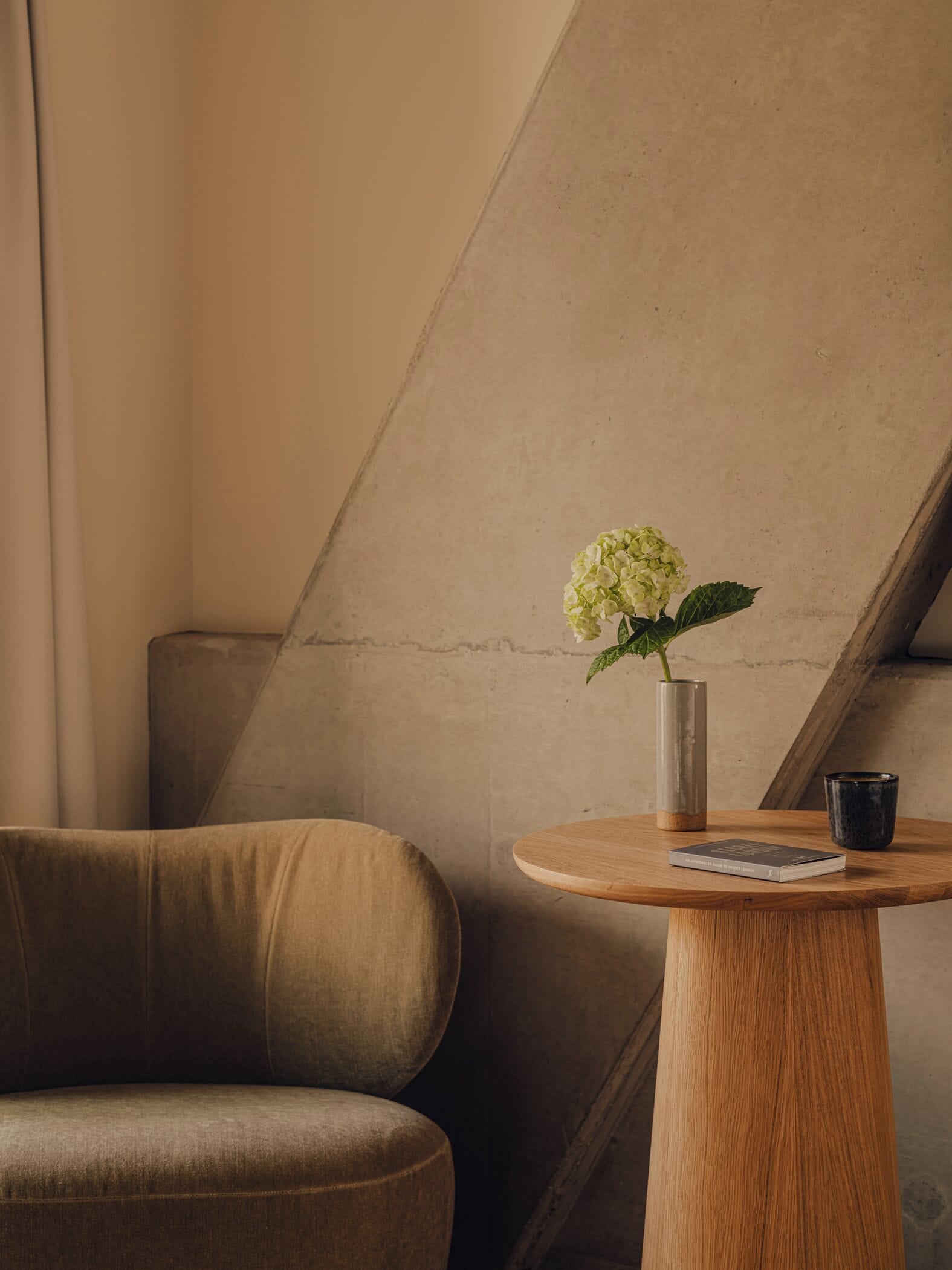 A corner of a room with a beige sofa, a round wooden table with a book and a vase of flowers, and a concrete wall.