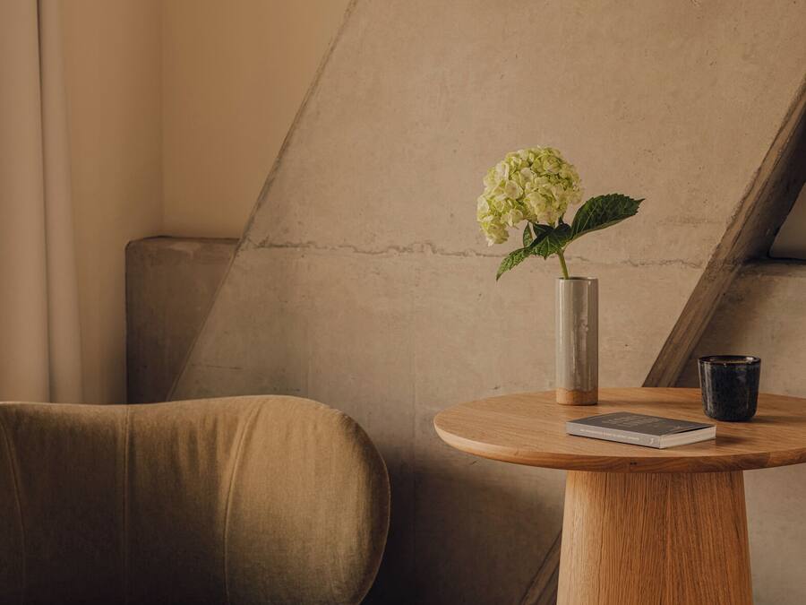 A corner of a room with a beige sofa, a round wooden table with a book and a vase of flowers, and a concrete wall.
