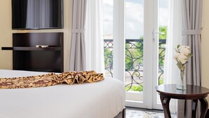A hotel room with a bed, a flat-screen TV mounted on the wall, a balcony with a wrought iron railing, and a vase with flowers on a small table. - Hoi An Canal House Riverside By Haviland (Da Nang)