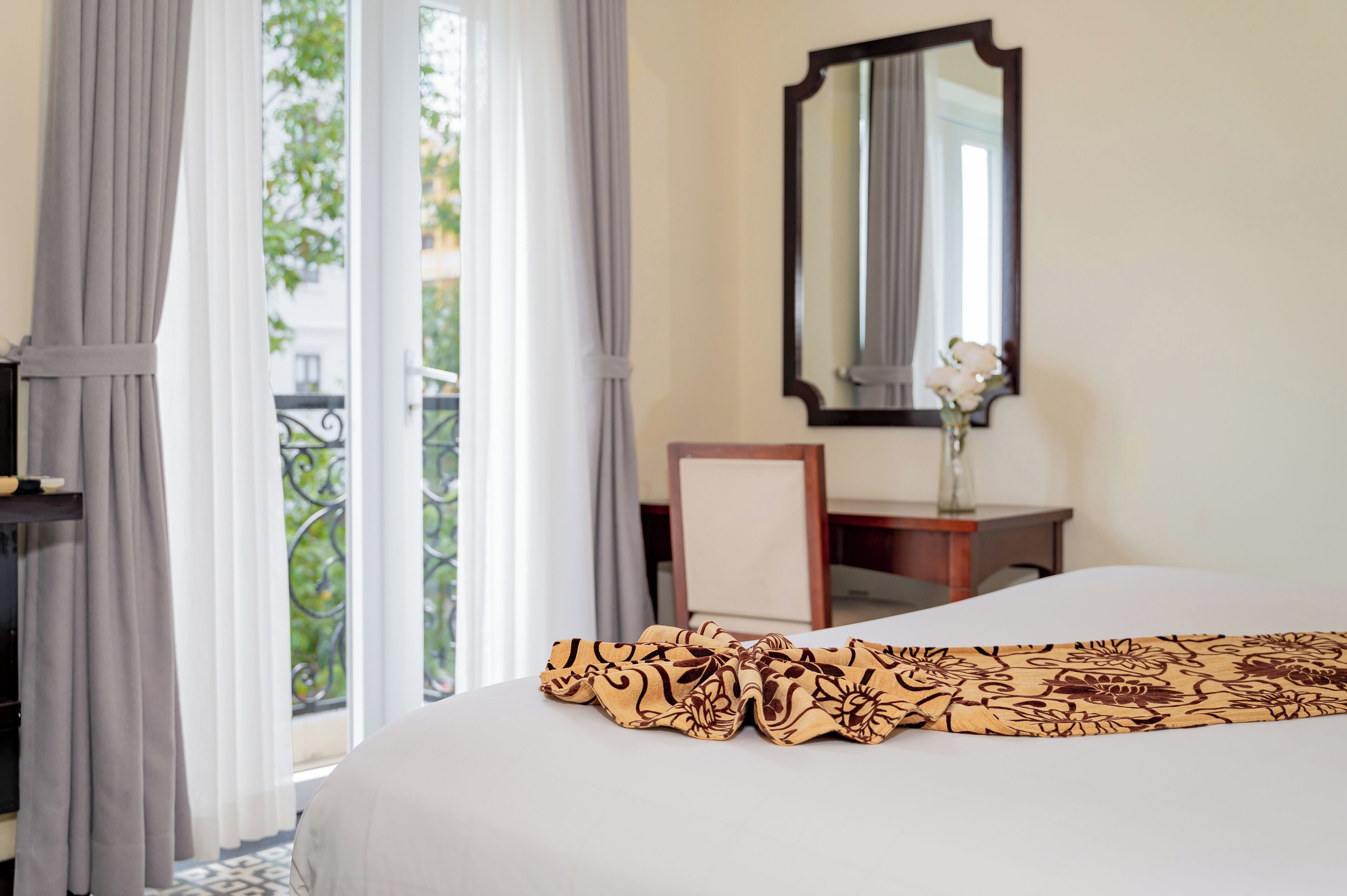 A hotel room with a bed, a desk, a chair, a mirror, and a view of greenery through the window.