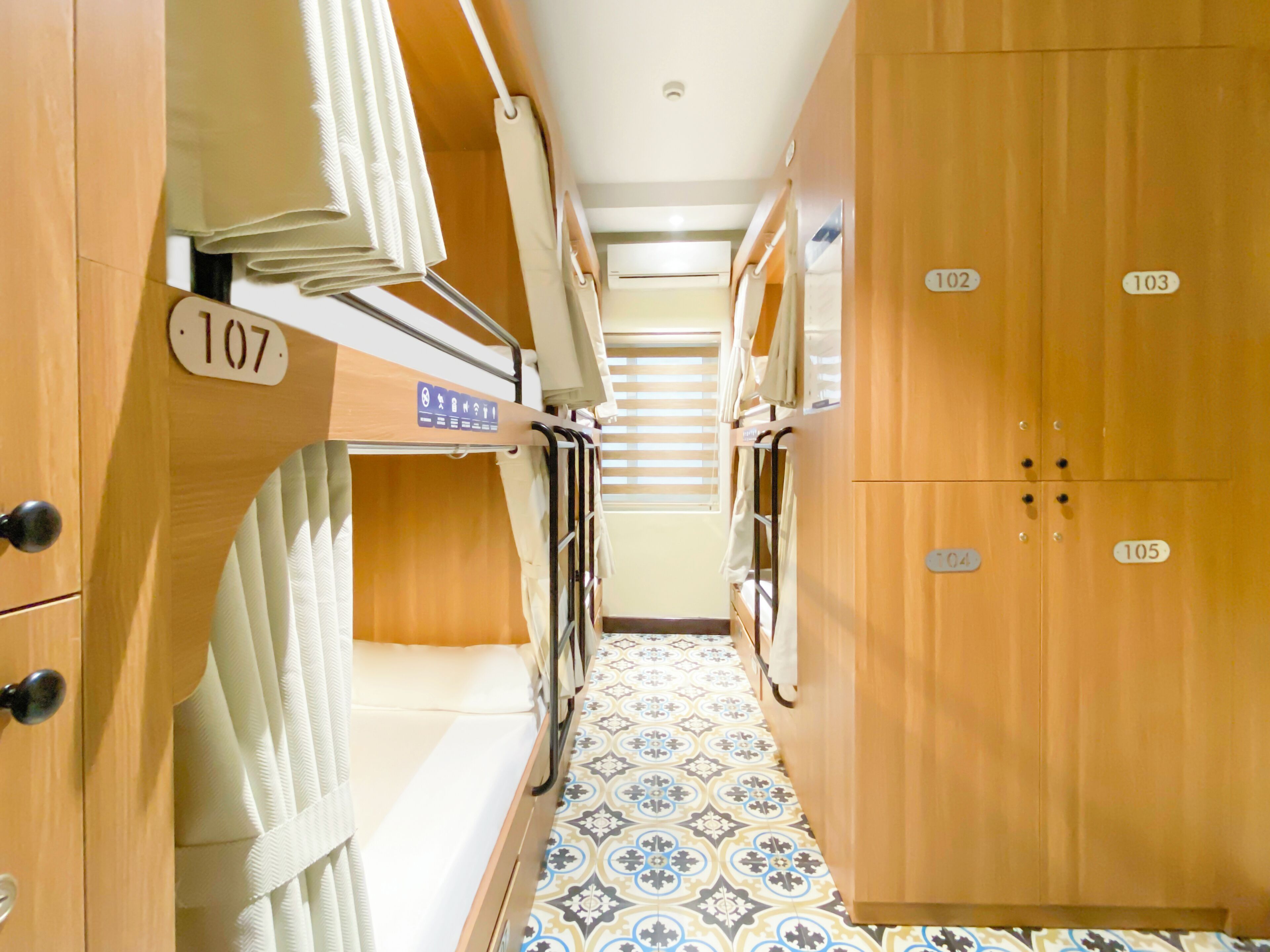 A narrow corridor with bunk beds, numbered doors, and patterned floor tiles.