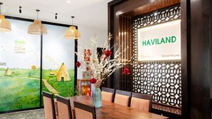 A dining area with a wooden table, chairs, and a vase with flowers. There is a wall with a geometric pattern and a sign that reads "HAVILAND Canal House". - Hoi An Canal House Riverside By Haviland (Da Nang)