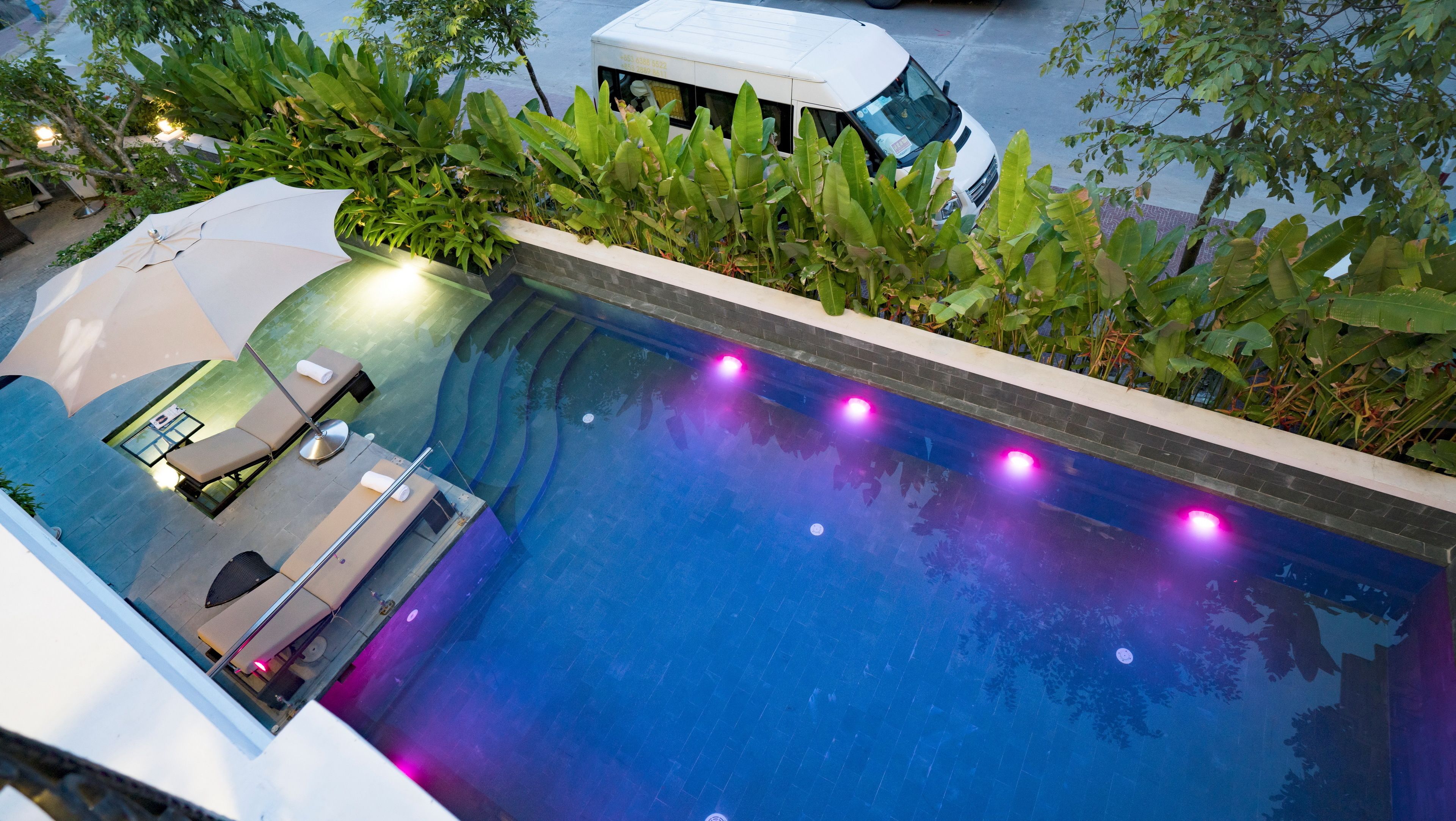 Una piscina al aire libre con un sillón de tomar el sol y una mesa, rodeada de abundante vegetación.