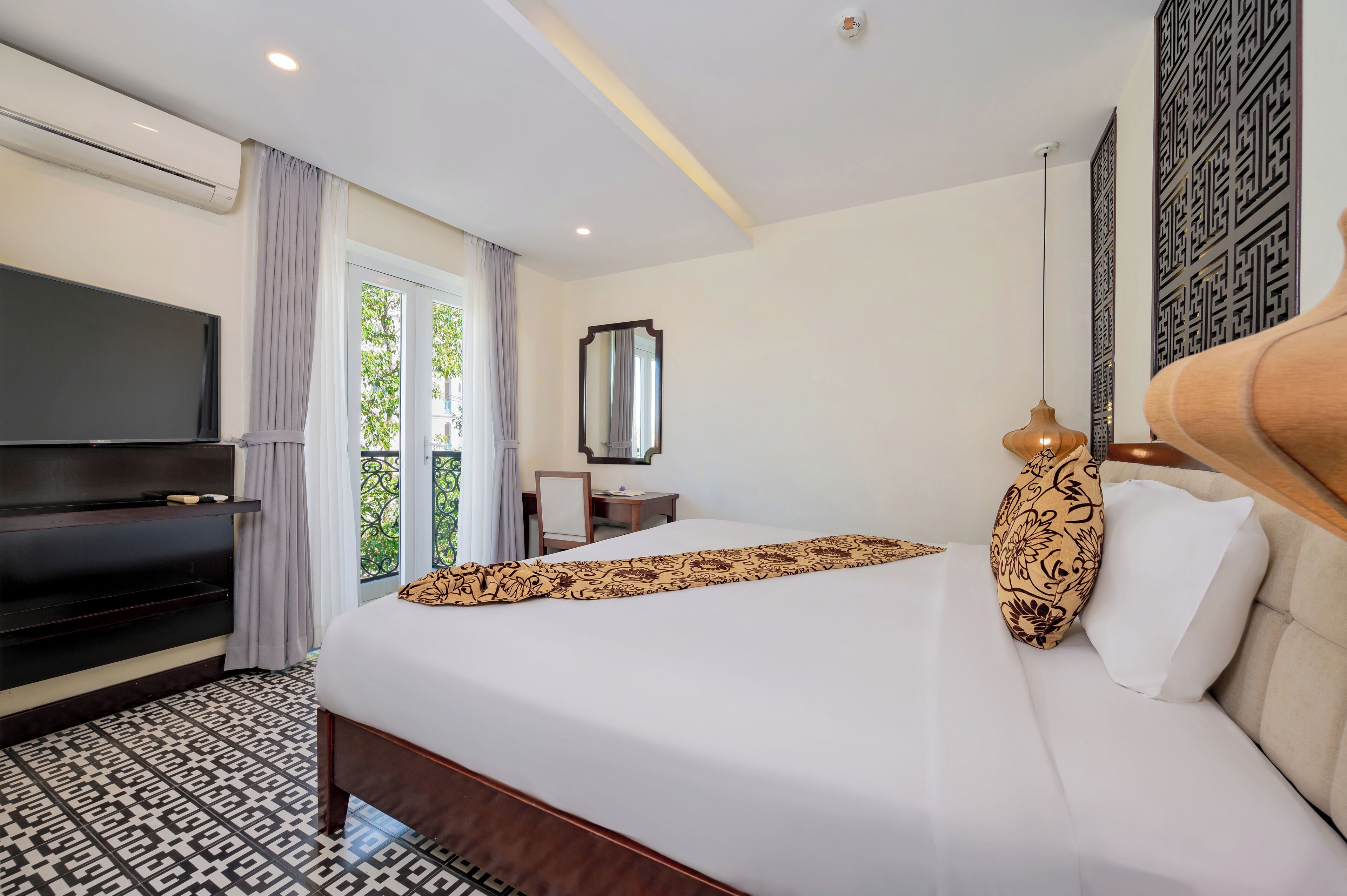A hotel room with a bed, a TV, a desk, and a view of greenery outside.