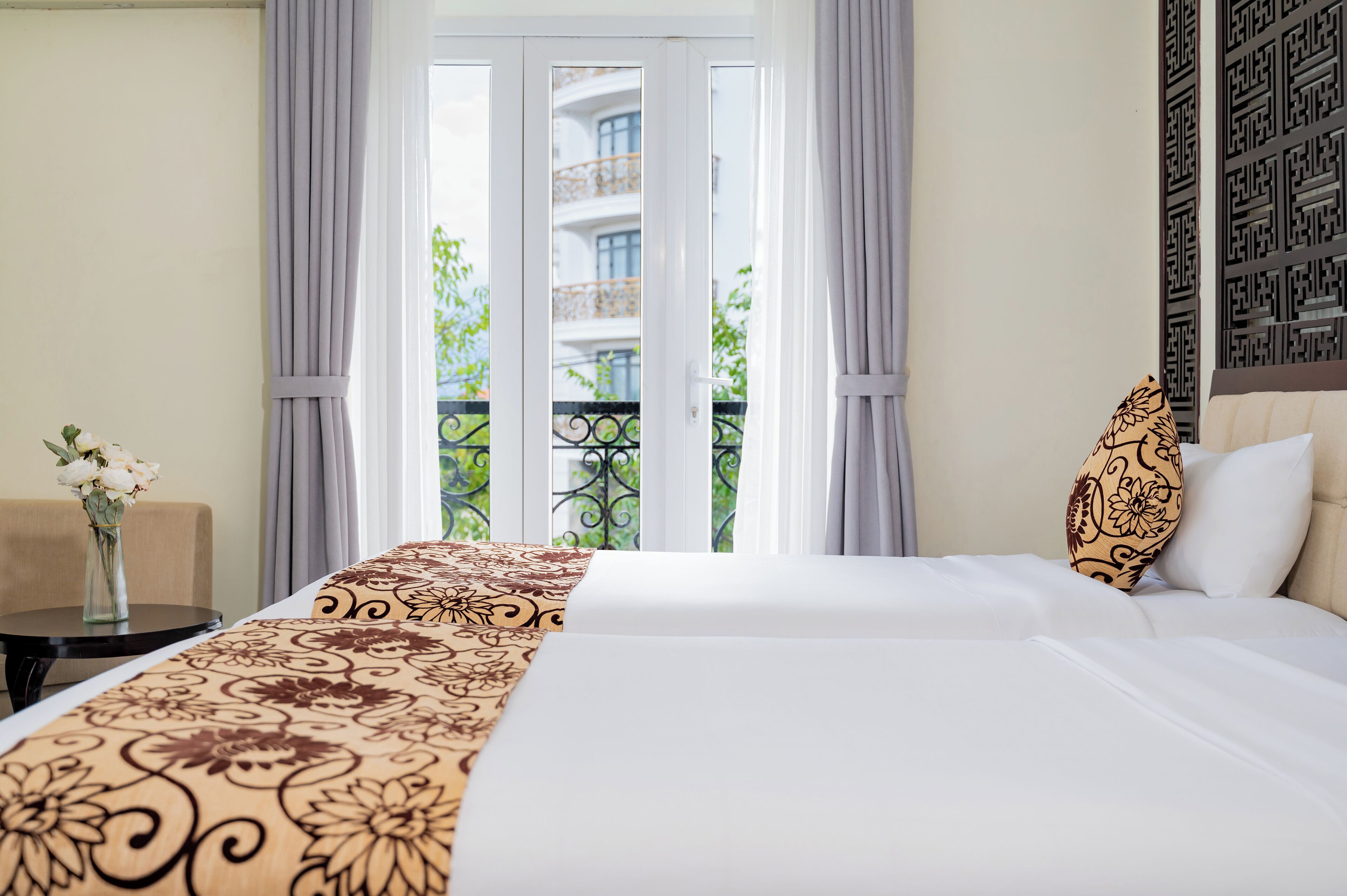 A hotel room with a bed, a small table with a vase of flowers, and a view of a balcony with greenery.