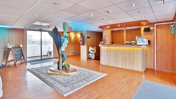 A hotel reception area with a wooden reception desk, a large decorative sculpture, and a view through the glass doors.