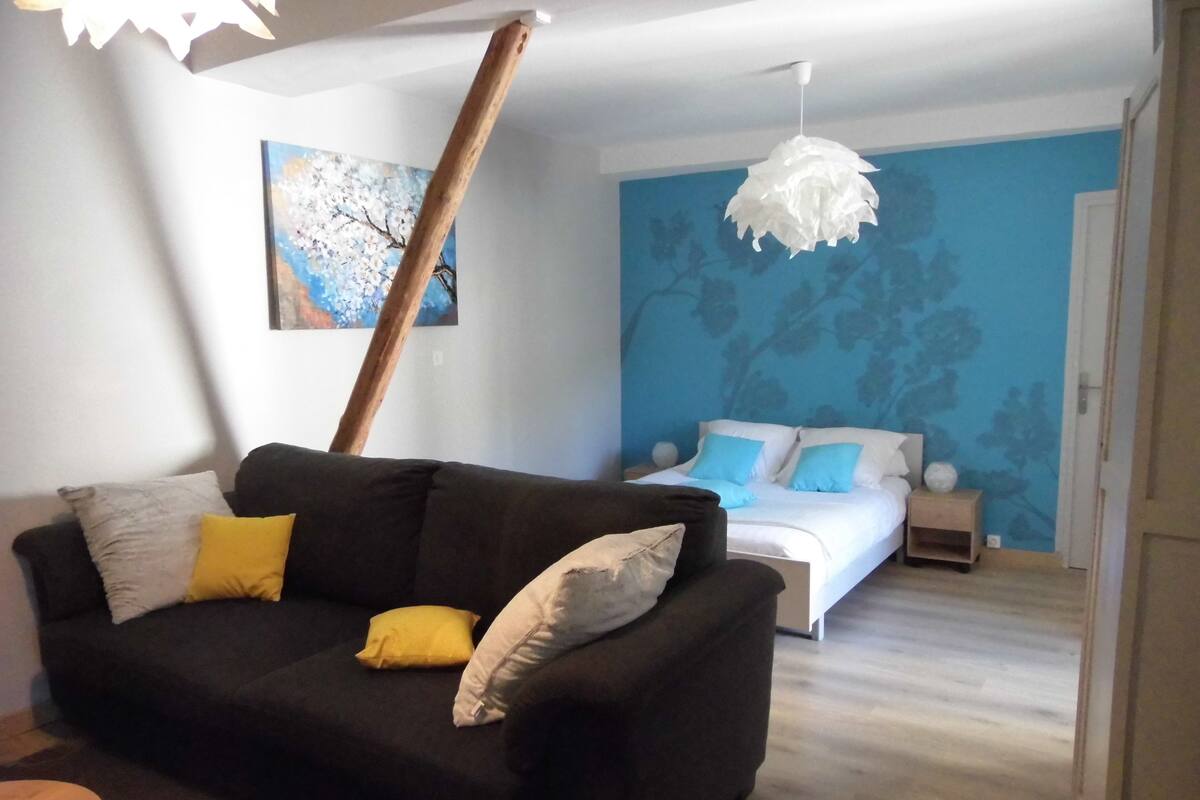 A modern room with a black sofa, a bed with blue and white bedding, a white hanging light fixture, and a blue wall with a tree pattern.