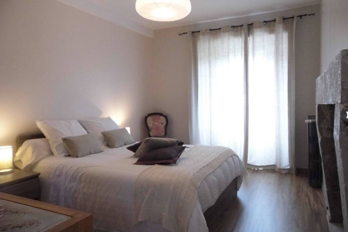 A bedroom with a bed, a nightstand, a chair, and a large window with curtains.
