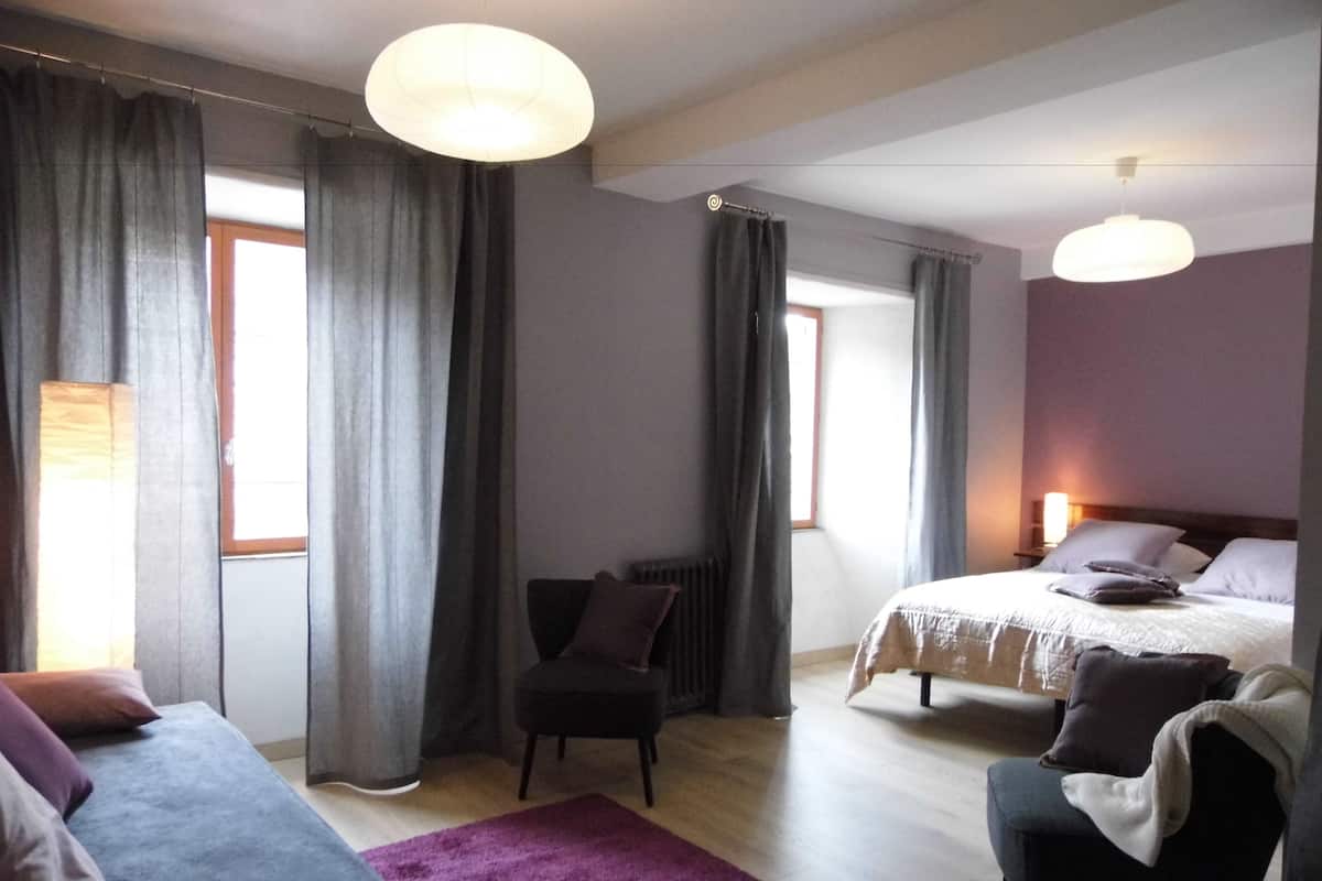 A hotel room with a bed, a chair, a sofa, and a large window with curtains.