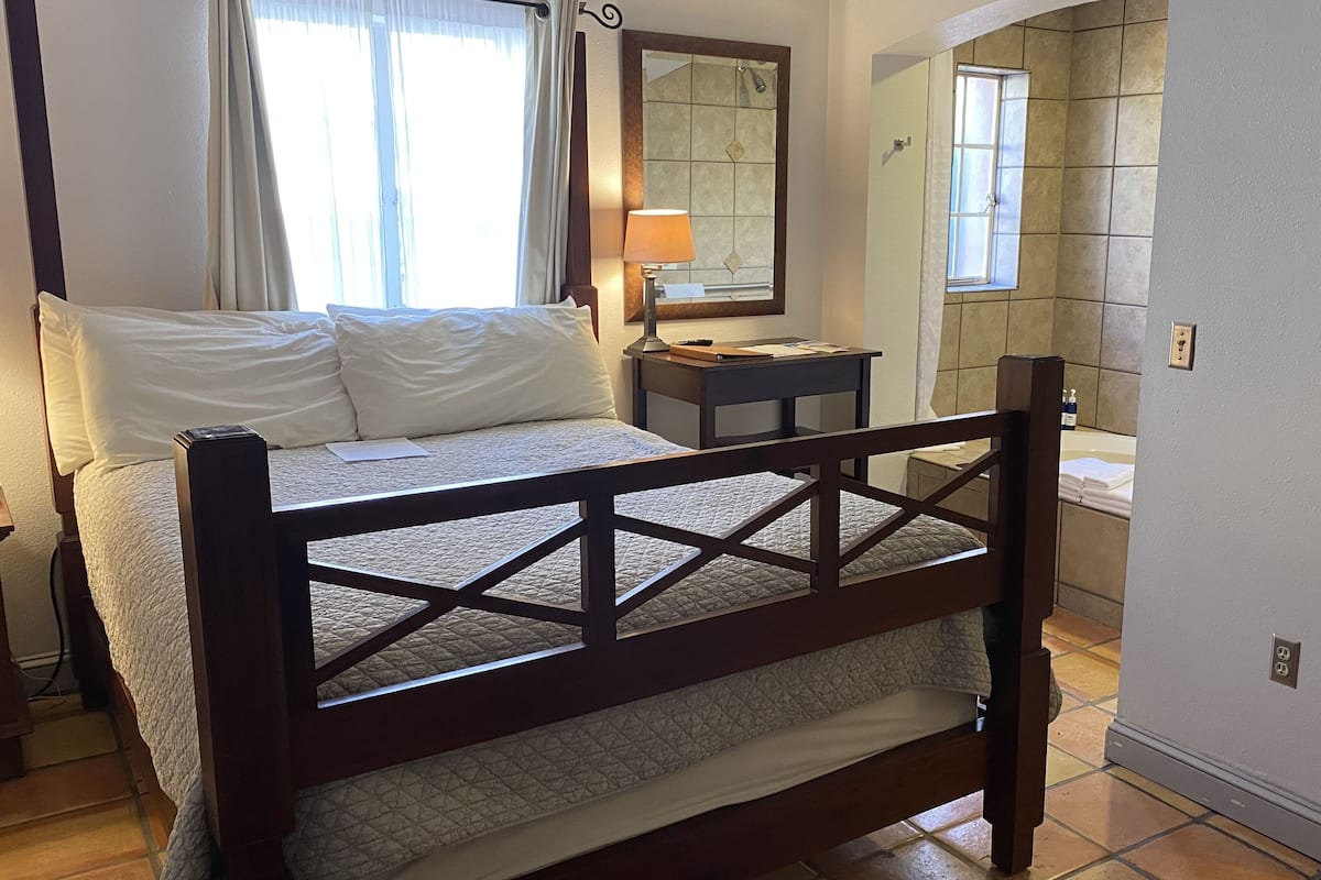 A hotel room with a wooden bed, a bedside table with a lamp, a mirror, and a view of the bathroom.