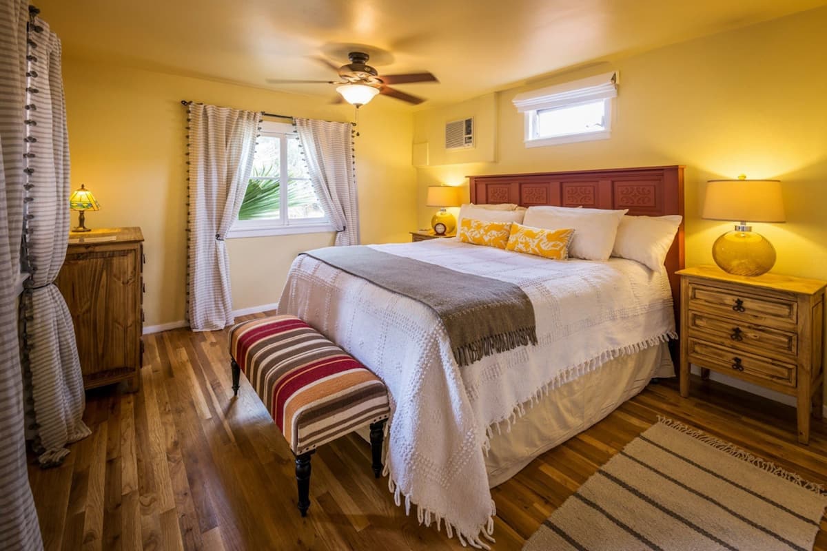 A bedroom with a bed, bedside lamps, a wooden bench, a ceiling fan, and a window with curtains.