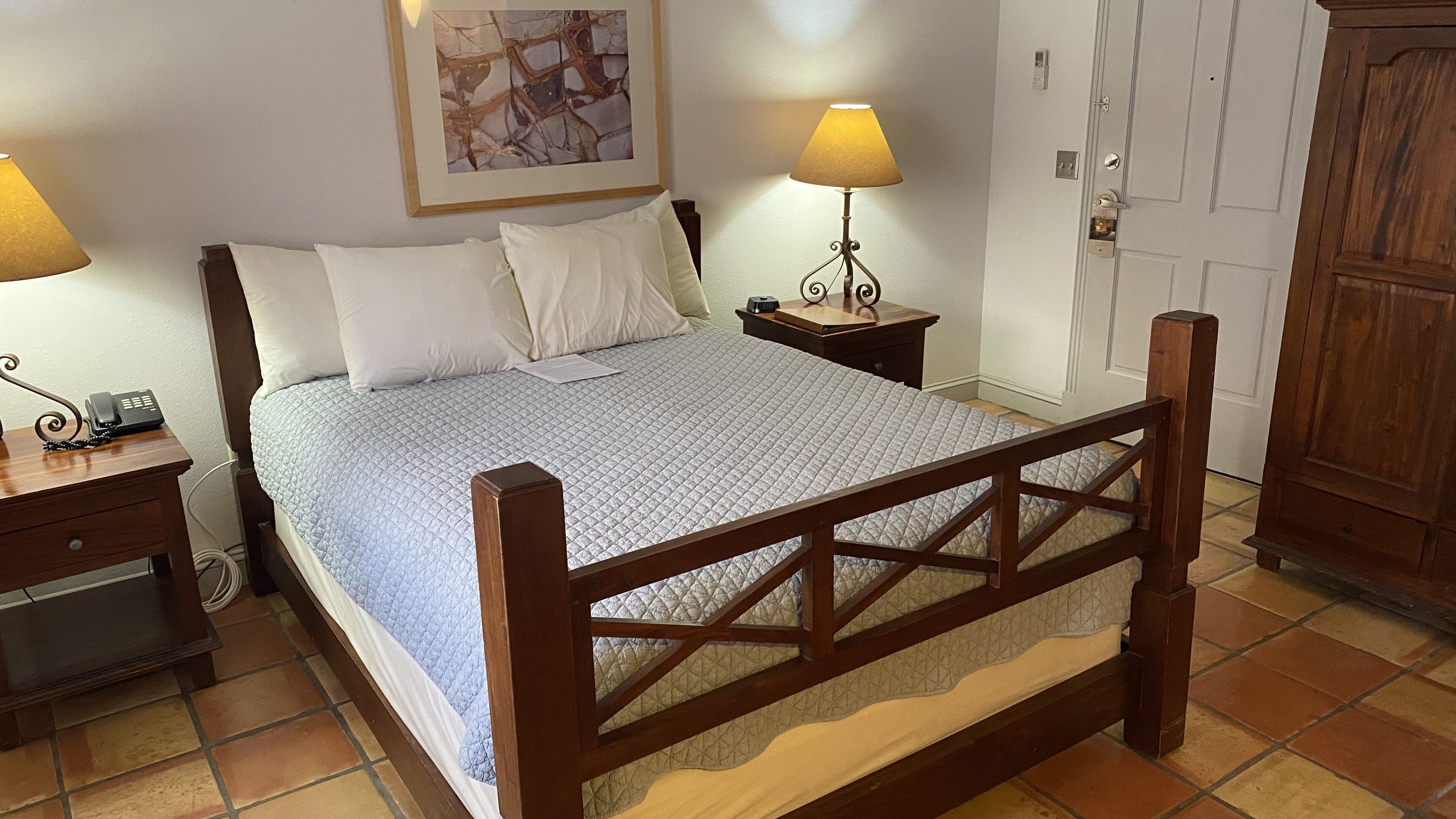 Ein Bett mit Holzbettgestell, einer gesteppten Matratze, zwei Nachttischlampen, einem Nachttisch, einem Telefon und einer Tür mit Griff.