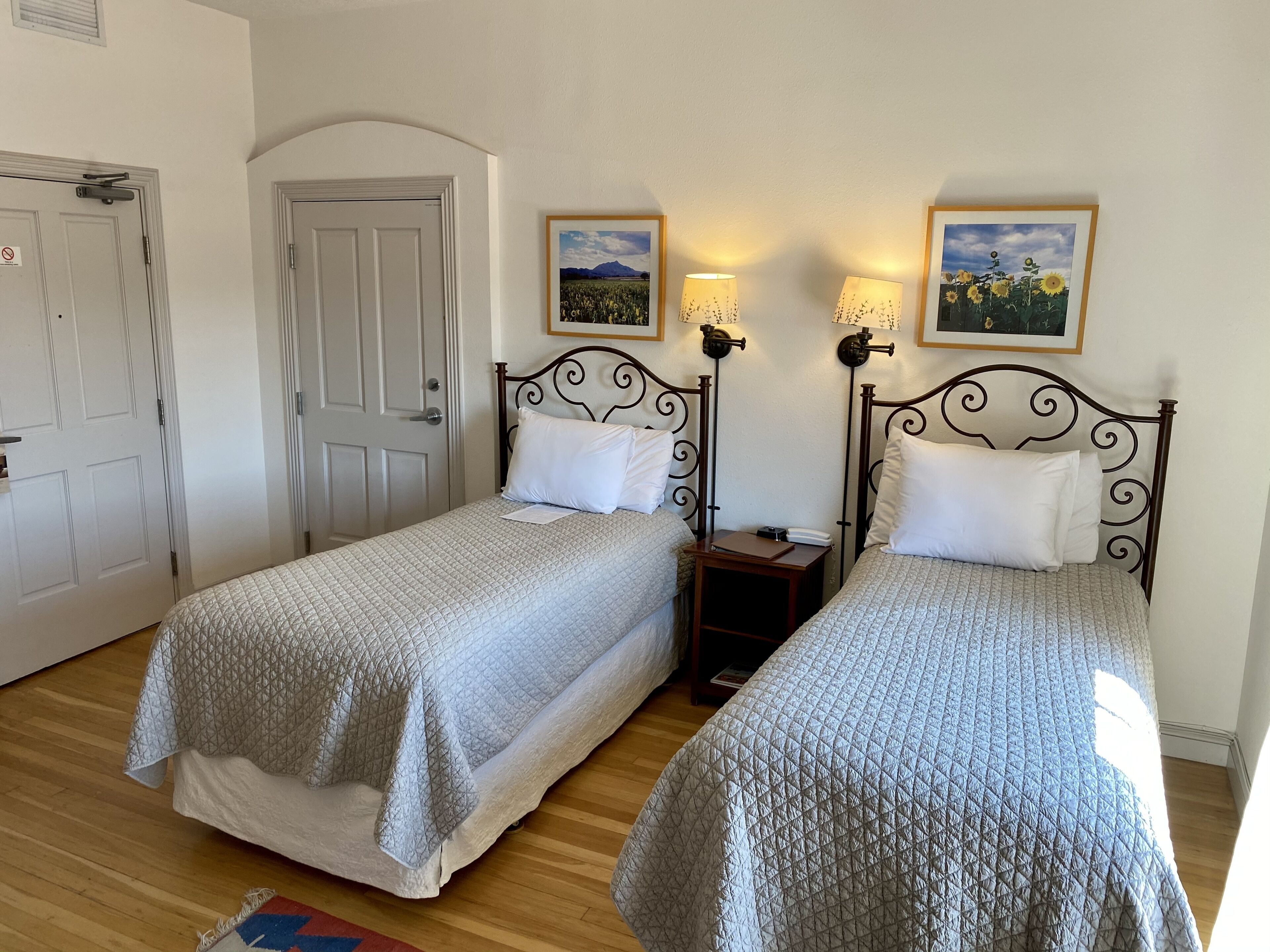 Two single beds with headboards, a wooden floor, a door, and two wall-mounted lamps with pictures.