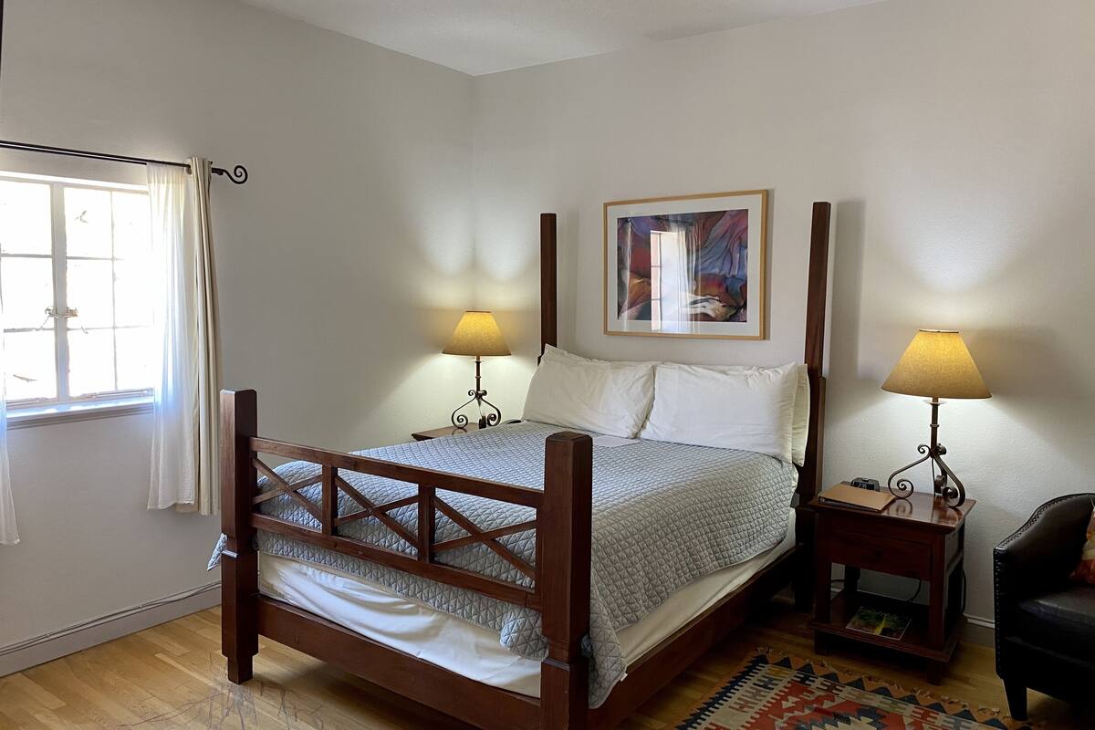 A bedroom with a wooden bed, two bedside lamps, a framed artwork on the wall, and a window with curtains.