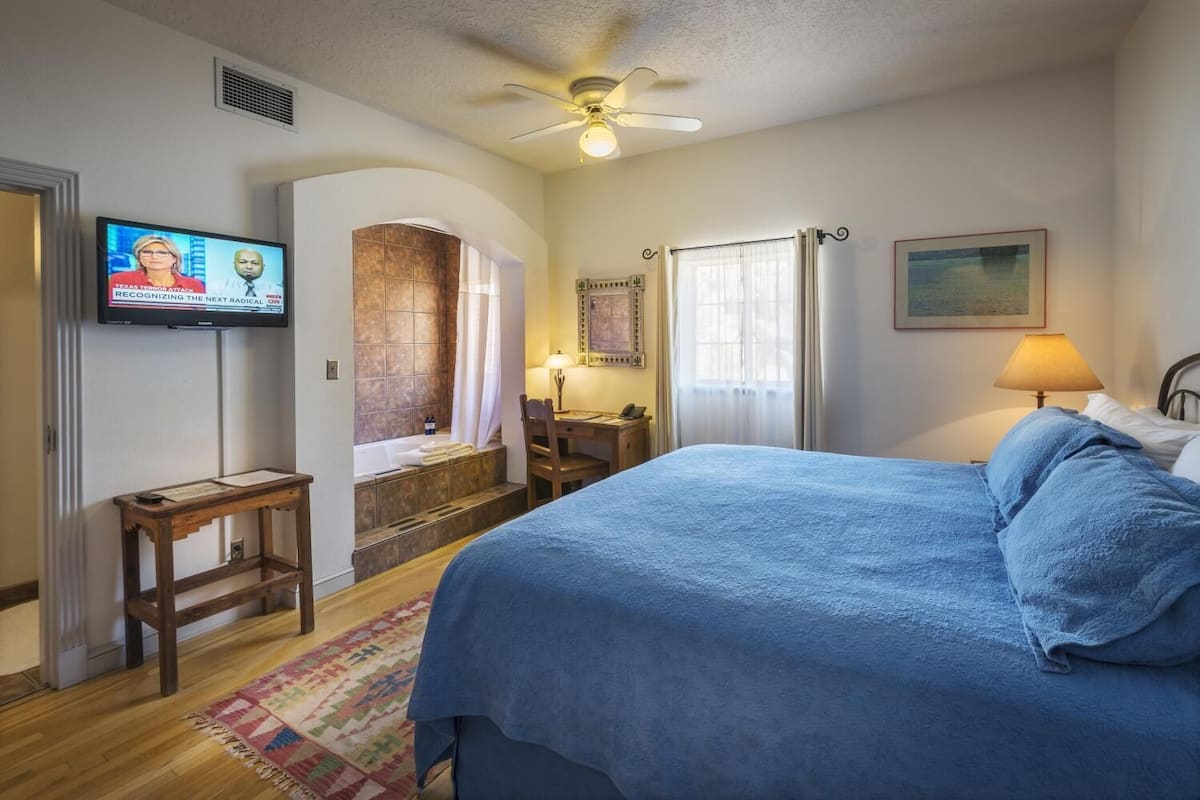A bedroom with a bed, a desk, a chair, a television, a window, and a bathroom with a bathtub.