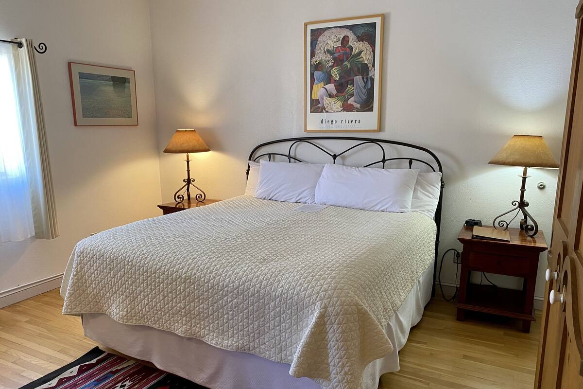 A neatly made bed with a quilted white duvet, two bedside lamps, a framed artwork above the bed, and a patterned rug on the floor.