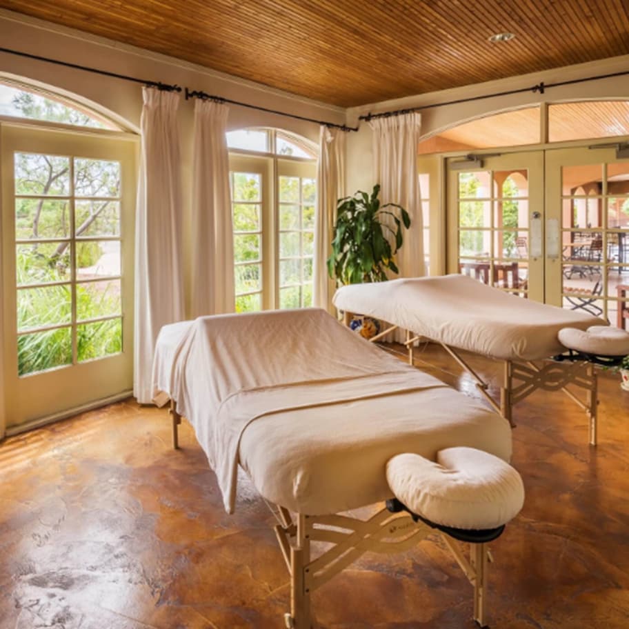 A room with two massage tables, large windows with curtains, a wooden ceiling, and a view of the outdoors.