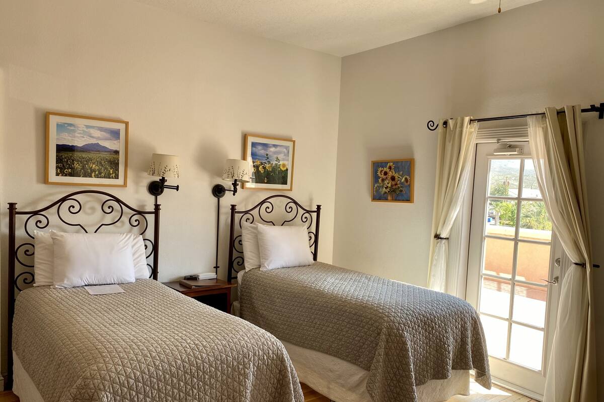 A room with two beds, each with a headboard, a nightstand, and a lamp. There are framed pictures on the wall, a window with curtains, and a door leading outside.