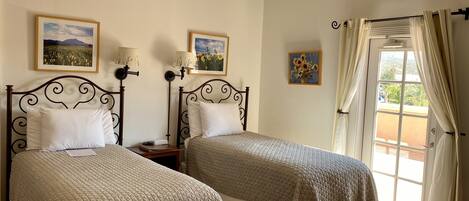 Una habitación con dos camas, cada una con cabecero, mesita de noche y lámpara. Se observan cuadros en la pared, una ventana con cortinas y una puerta que da al exterior.