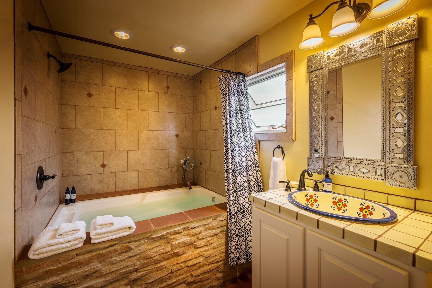 Baño con ducha de azulejos de piedra, bañera, lavabo con espejo decorativo y toallas sobre el suelo.