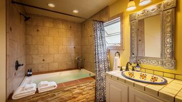 Baño con ducha de azulejos de piedra, bañera, lavabo con espejo decorativo y toallas sobre el suelo.