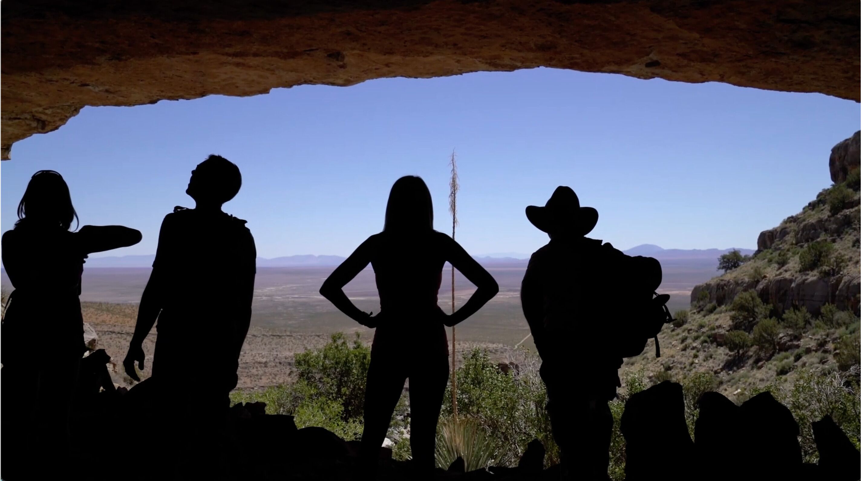 Un grupo de personas siluetas contra un cielo despejado, de pie en un entorno natural con paisaje rocoso y vegetación escasa.